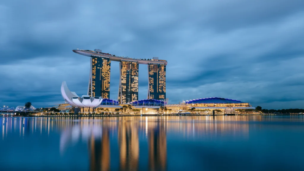 Night view of Marina Bay Sands hotel and the ArtScience Museum in Singapore, with illuminated buildings reflecting on calm water under a cloudy sky.