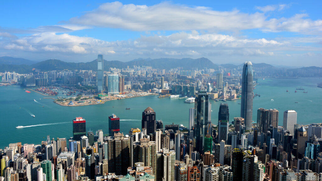 A wide view of Hong Kong’s skyline showing tall skyscrapers, Victoria Harbour, and mountains in the background under a blue sky with scattered clouds.