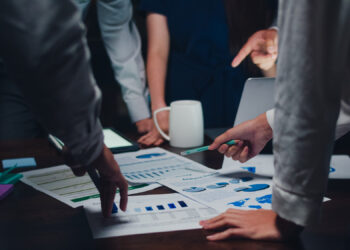 Colleagues gather around a table, scrutinizing charts and graphs with intense focus. Hands gesture at various documents, while a white mug and an open laptop signal a strategic discussion. The scene suggests a collaborative work meeting centered on the development of an innovative business plan.