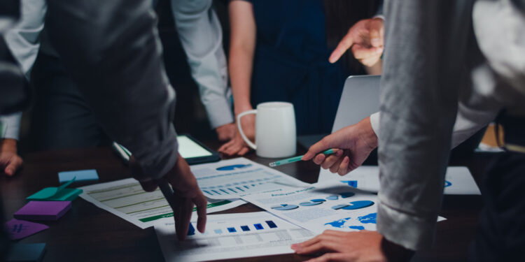 Colleagues gather around a table, scrutinizing charts and graphs with intense focus. Hands gesture at various documents, while a white mug and an open laptop signal a strategic discussion. The scene suggests a collaborative work meeting centered on the development of an innovative business plan.