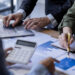 Three people collaborate at a desk with charts, a laptop, a calculator, and a notepad. One person types, another writes notes, and another points at a graph, suggesting a business or financial discussion.
