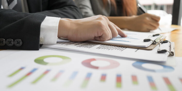 Close-up of a person in business attire pointing at a graph on a clipboard, with colorful charts and graphs on paper in the foreground and another person writing in the background.