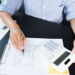 A person in a striped shirt is using a calculator while reviewing financial documents and charts at a desk.