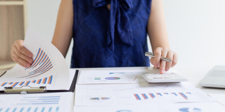 A person in a navy blue blouse is reviewing financial charts and graphs on paper, using a calculator at a desk filled with documents and a laptop.