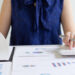 A person in a navy blue blouse is reviewing financial charts and graphs on paper, using a calculator at a desk filled with documents and a laptop.