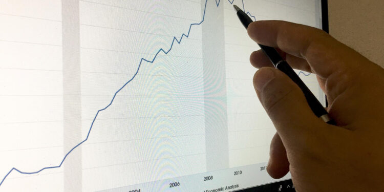 A hand holding a pen points at a line graph displayed on a computer monitor, showing trends from 2000 to 2015 with marked periods of recession in the background.