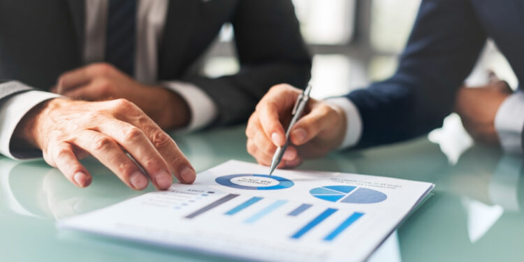 Two people in business suits review financial charts and graphs on a document, discussing data and pointing at a pie chart with a pen while seated at a glass table in a bright office.