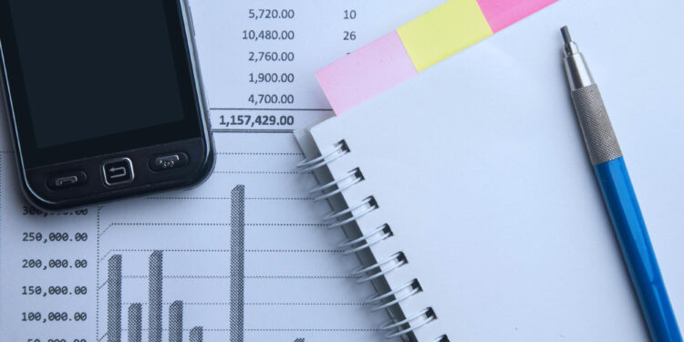 A smartphone, sticky notes, a pen, a spiral notebook, and financial documents with charts and numbers are arranged on a desk, suggesting a workspace focused on data analysis or accounting.
