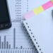 A smartphone, sticky notes, a pen, a spiral notebook, and financial documents with charts and numbers are arranged on a desk, suggesting a workspace focused on data analysis or accounting.