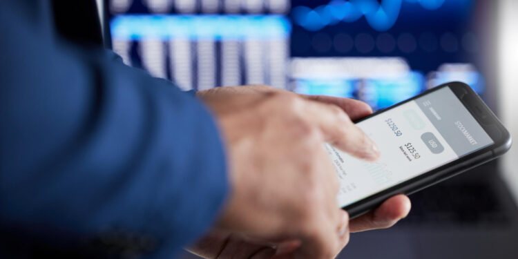 A person in a suit uses a smartphone with a financial app open, showing account balances. In the blurred background, a computer screen displays financial charts and data.