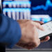 A person in a suit uses a smartphone with a financial app open, showing account balances. In the blurred background, a computer screen displays financial charts and data.