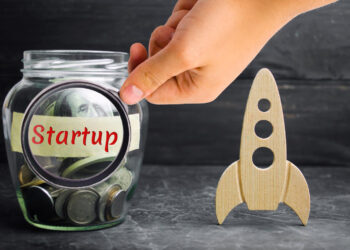 A hand places a coin into a glass jar labeled "Startup" filled with coins and cash, next to a wooden rocket figurine, symbolizing investment in a business startup.