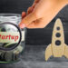 A hand places a coin into a glass jar labeled "Startup" filled with coins and cash, next to a wooden rocket figurine, symbolizing investment in a business startup.