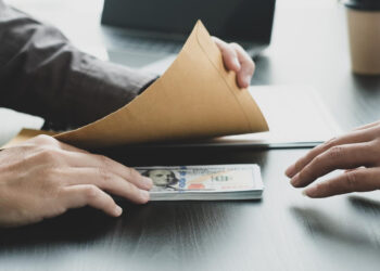 Two people at a table exchange a stack of US hundred-dollar bills inside a brown envelope, suggesting a secretive financial transaction. Laptops and a coffee cup are visible in the background.