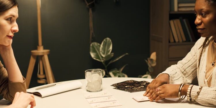 Two women sit at a round table. One is laying tarot cards in front of the other. A drink with ice and a notebook are on the table. The setting is warmly lit and calm, with plants and shelves in the background.