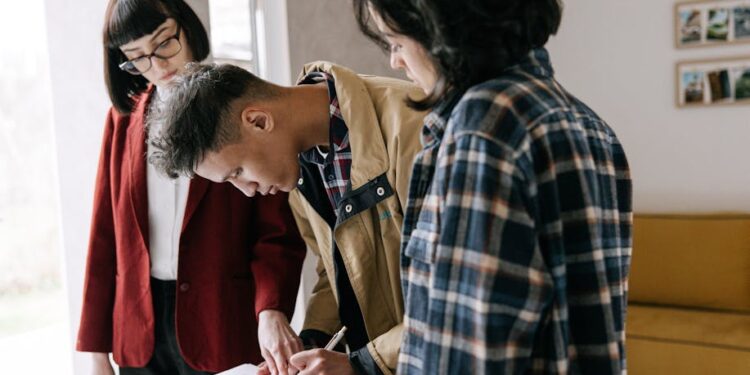 Three people stand together indoors as one person signs a document on a clipboard, while another points at the paper and the third observes. They appear focused and engaged in the task.
