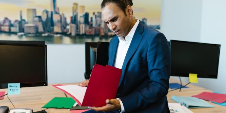A man in a blue suit sits on a desk in an office, looking at papers in a red folder. Two computer monitors, files, and sticky notes are on the desk. A cityscape photo hangs on the wall behind him.