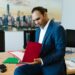 A man in a blue suit sits on a desk in an office, looking at papers in a red folder. Two computer monitors, files, and sticky notes are on the desk. A cityscape photo hangs on the wall behind him.